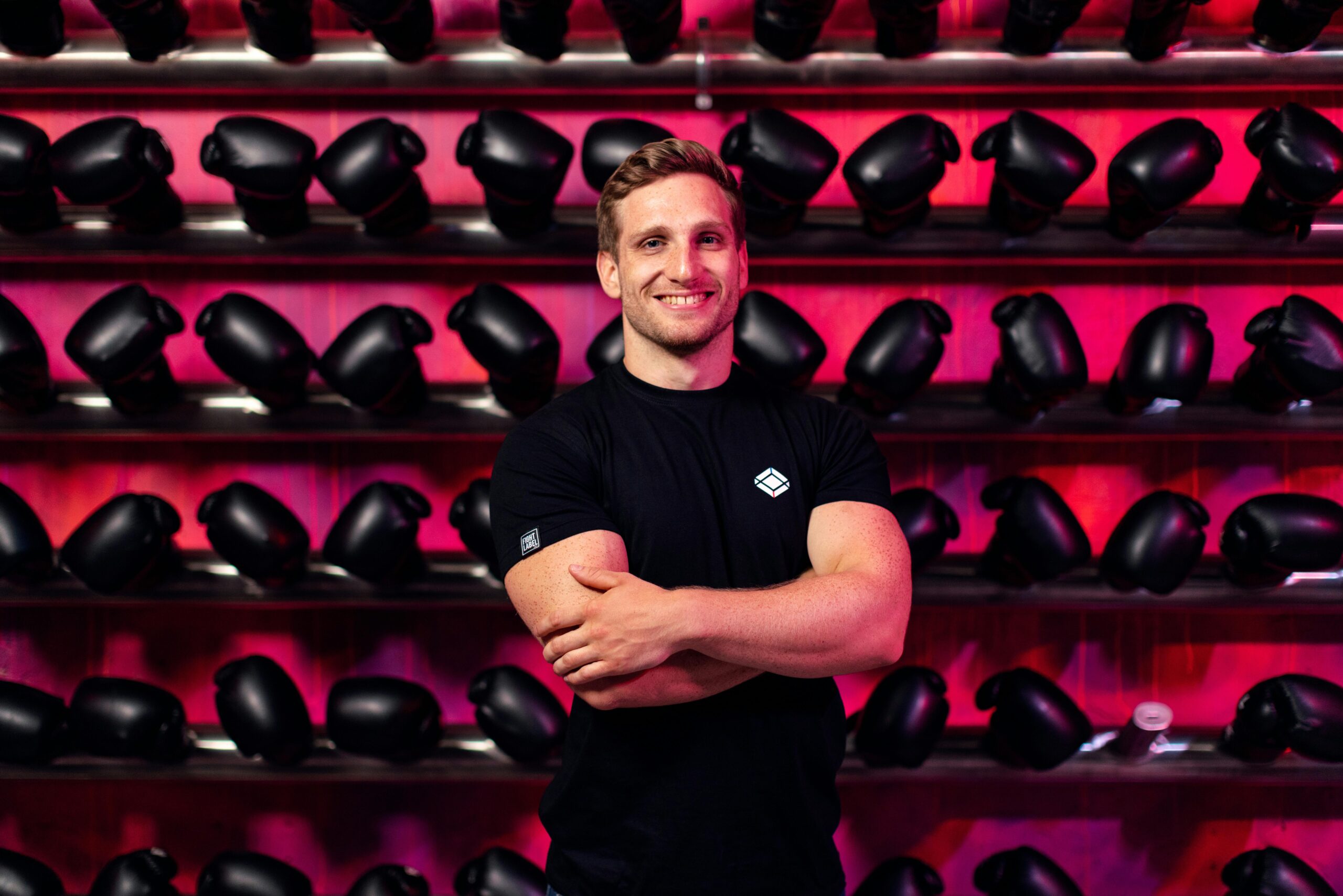 A smiling man in a gym stands confidently in front of a wall of boxing gloves.